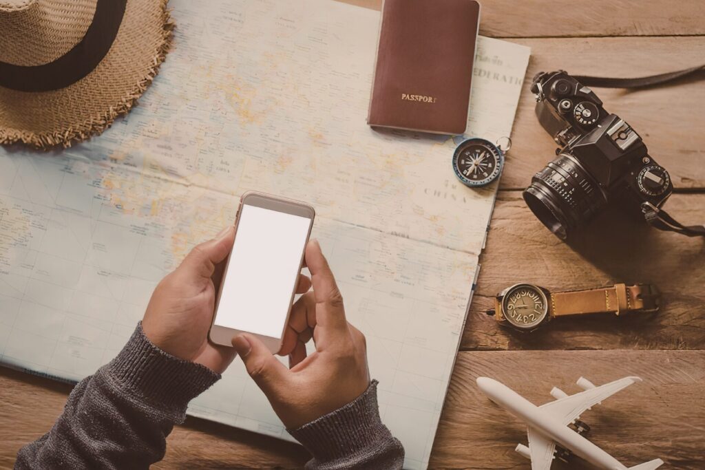 Hands holding a smartphone over a travel map with a passport, camera and compass